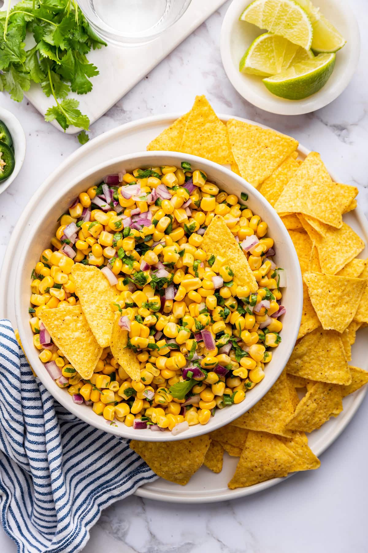 Corn mixed with red onion, cilantro and jalapeño in a white bowl served with tortilla chips.