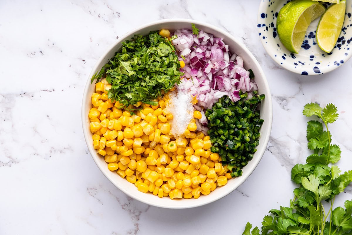 Ingredients for the corn salsa: corn, red onion, cilantro, jalapeño, salt and lime juice in a white serving bowl.