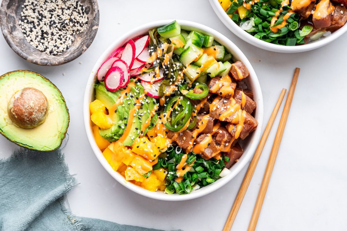 A serving of a poke bowl with fresh vegetables, tuna, sesame seeds, and a drizzle of a mayo based sauce.