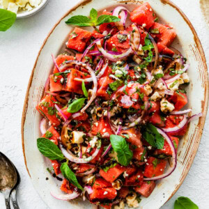 A large dish with a watermelon salad that is garnished with fresh basil, feta cheese, balsamic vinegar, and red onions.
