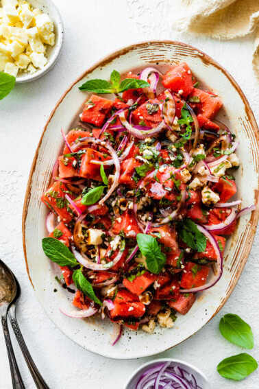 A large dish with a watermelon salad that is garnished with fresh basil, feta cheese, balsamic vinegar, and red onions.