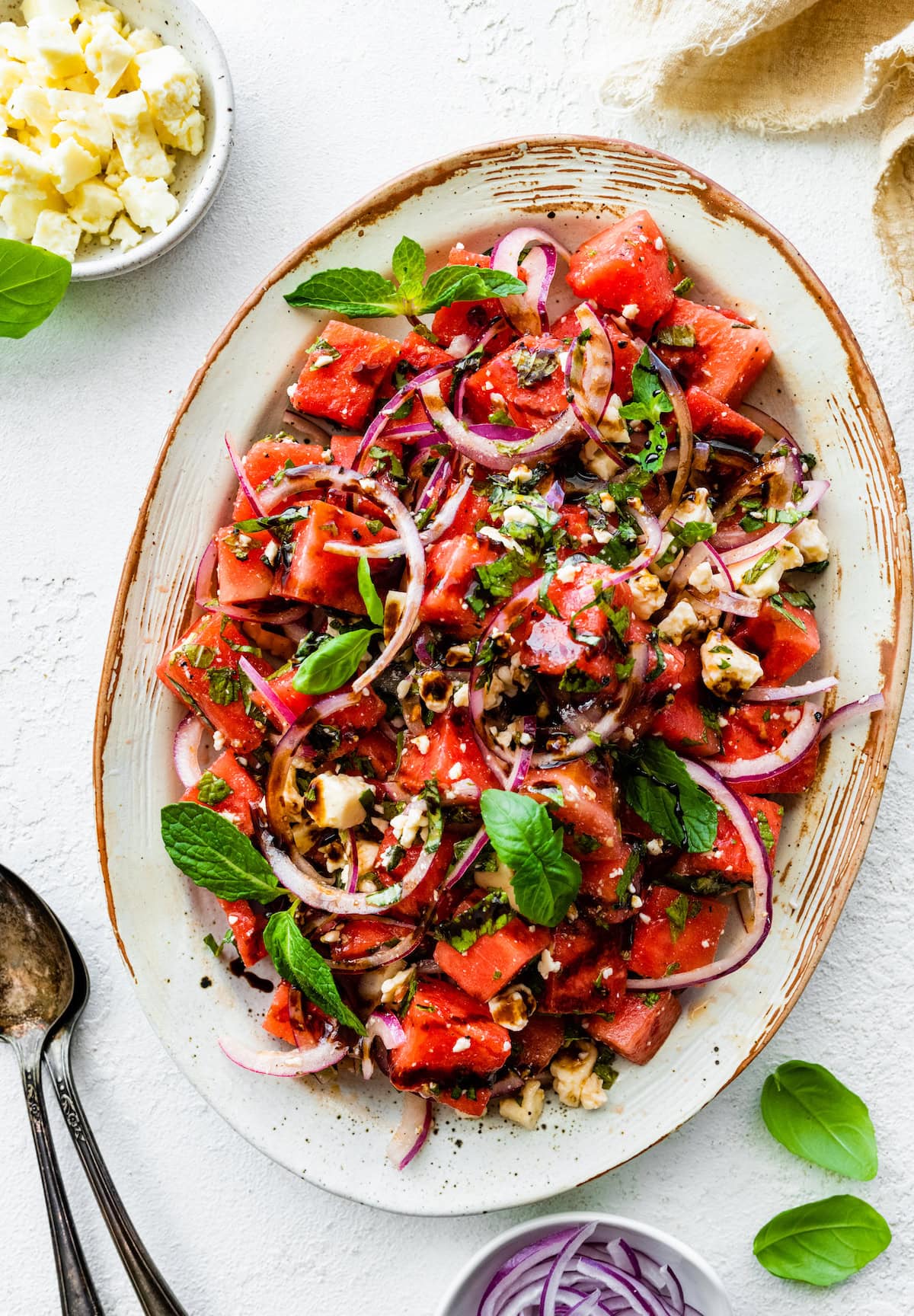A large dish with a watermelon salad that is garnished with fresh basil, feta cheese, balsamic vinegar, and red onions.