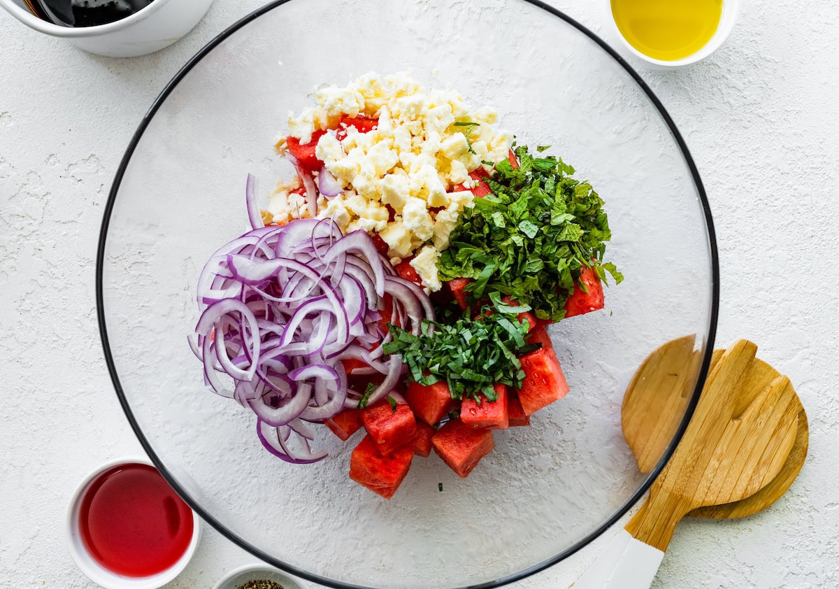 A large glass bowl with red onion, basil, feta cheese, and watermelon cubes.