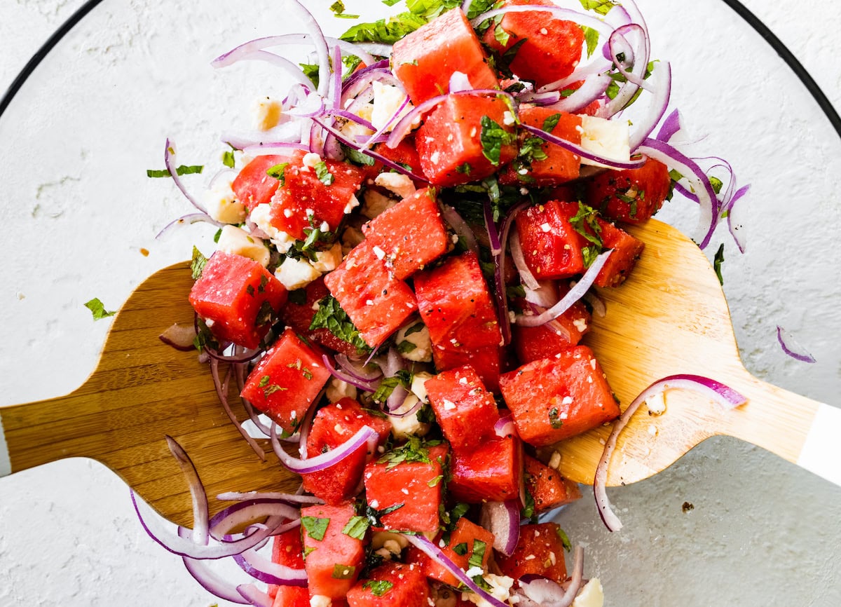 Two wooden serving spatulas mix together watermelon salad in a large glass bowl.