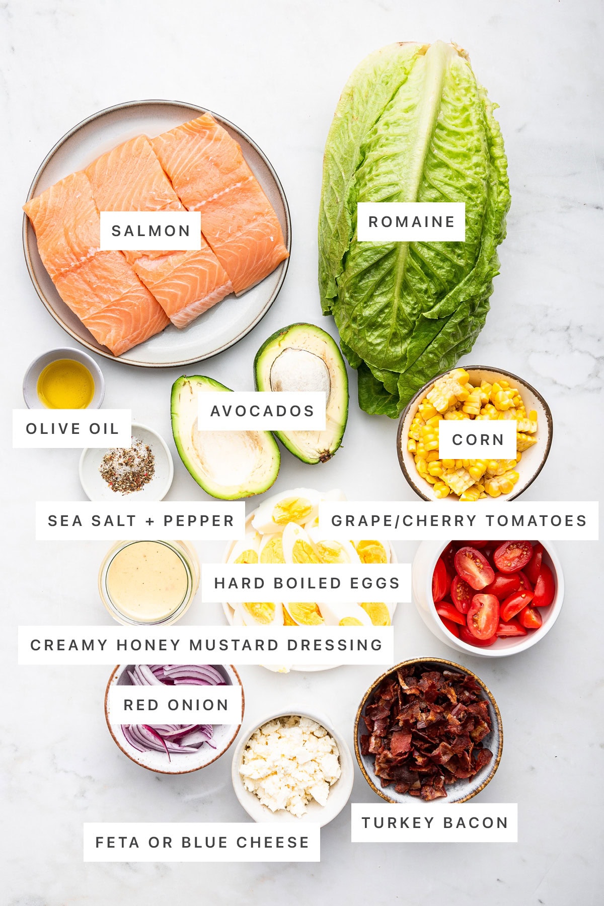 Ingredients measured out to make a salmon cobb salad: salmon, romaine, olive oil, salt, pepper, avocados, corn, cherry tomatoes, hard boiled eggs, creamy honey mustard dressing, red onion, feta and turkey bacon.