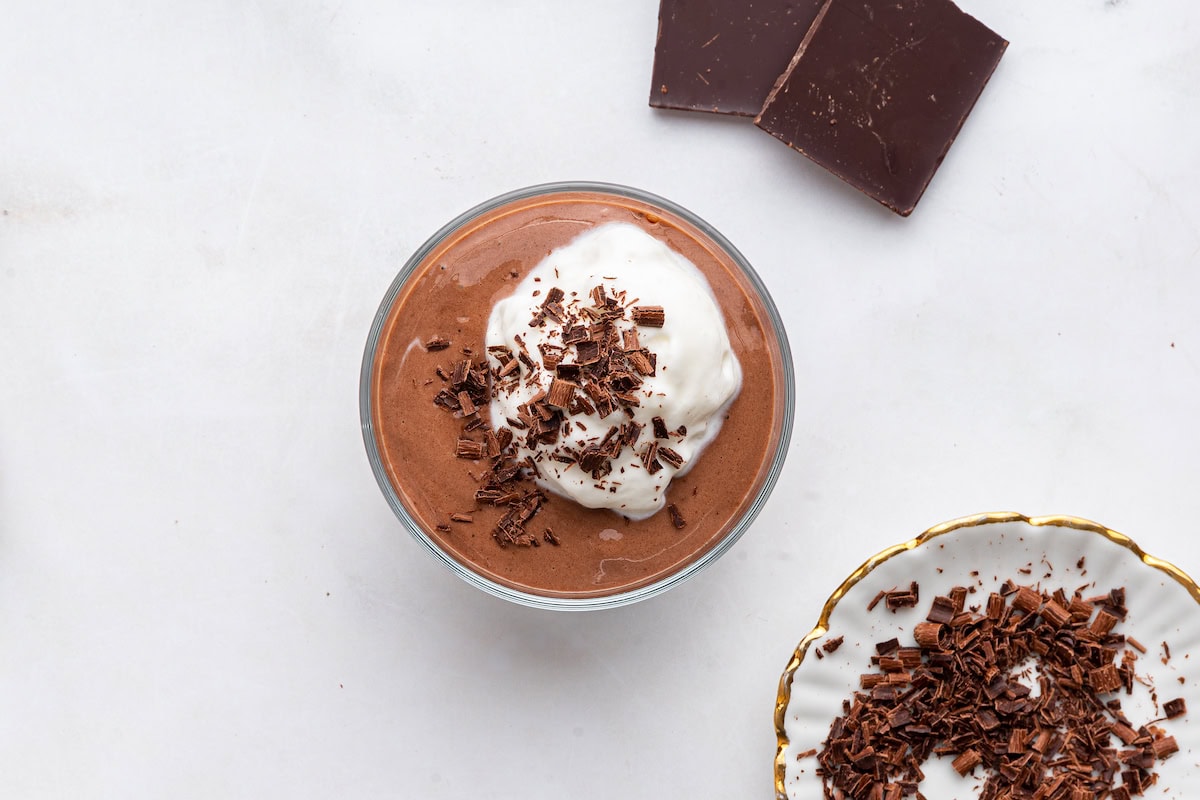 Chocolate protein pudding in a glass cup that is topped with whipped cream and shaved chocolate with a chocolate bar to the side.