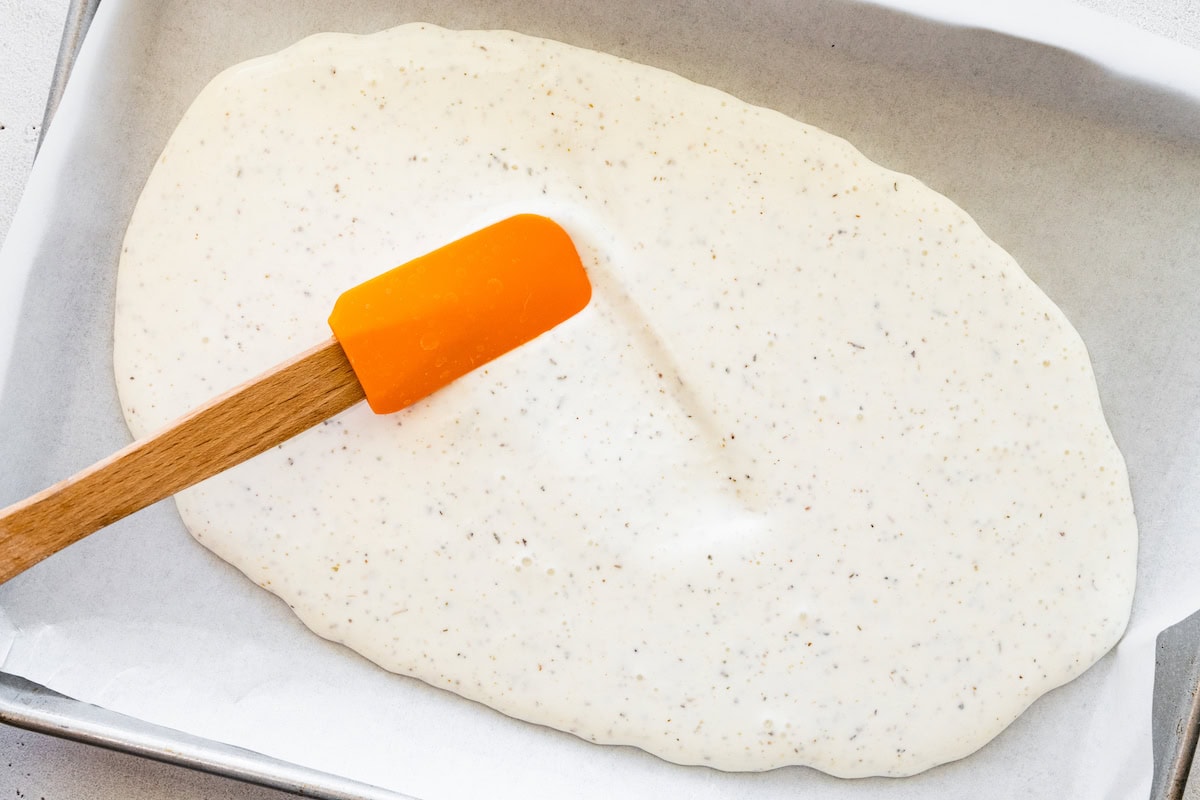 A spatula spreading the cottage cheese wrap mixture in a baking tray on parchment paper.