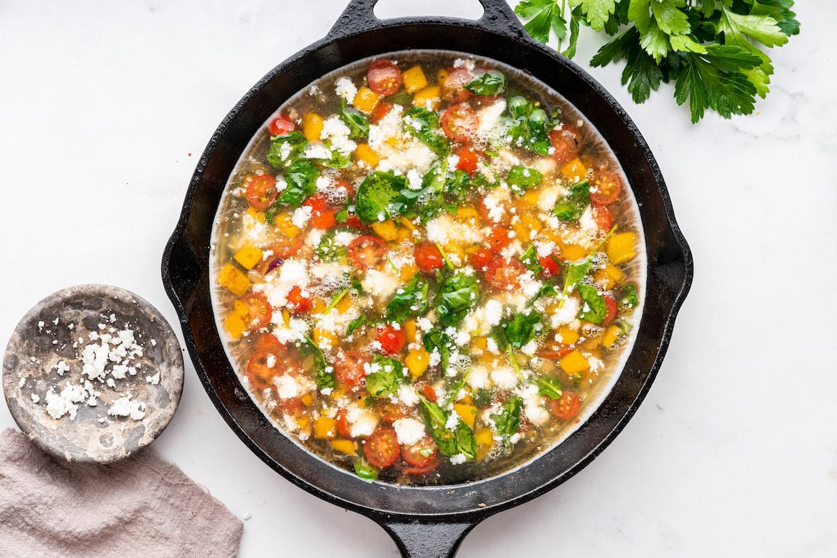Crumbled feta cheese is added to a cast iron skillet with cherry tomatoes, spinach, bell pepper, and red onion.