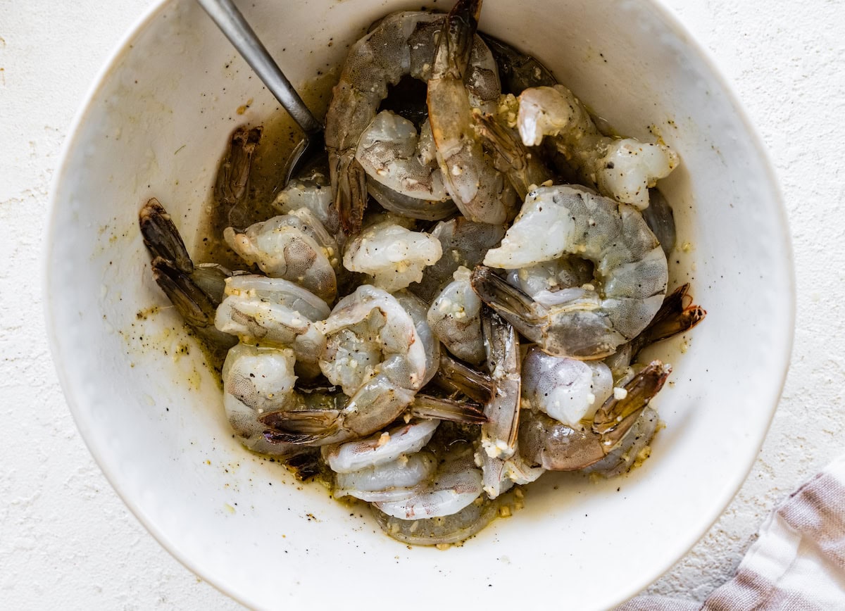 Raw shrimp in a medium mixing bowl with garlic, lemon zest, lemon juice, oil, salt and pepper.