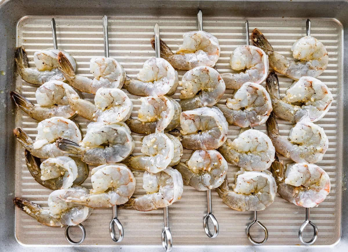 Raw shrimp on metal skewers on a baking tray.