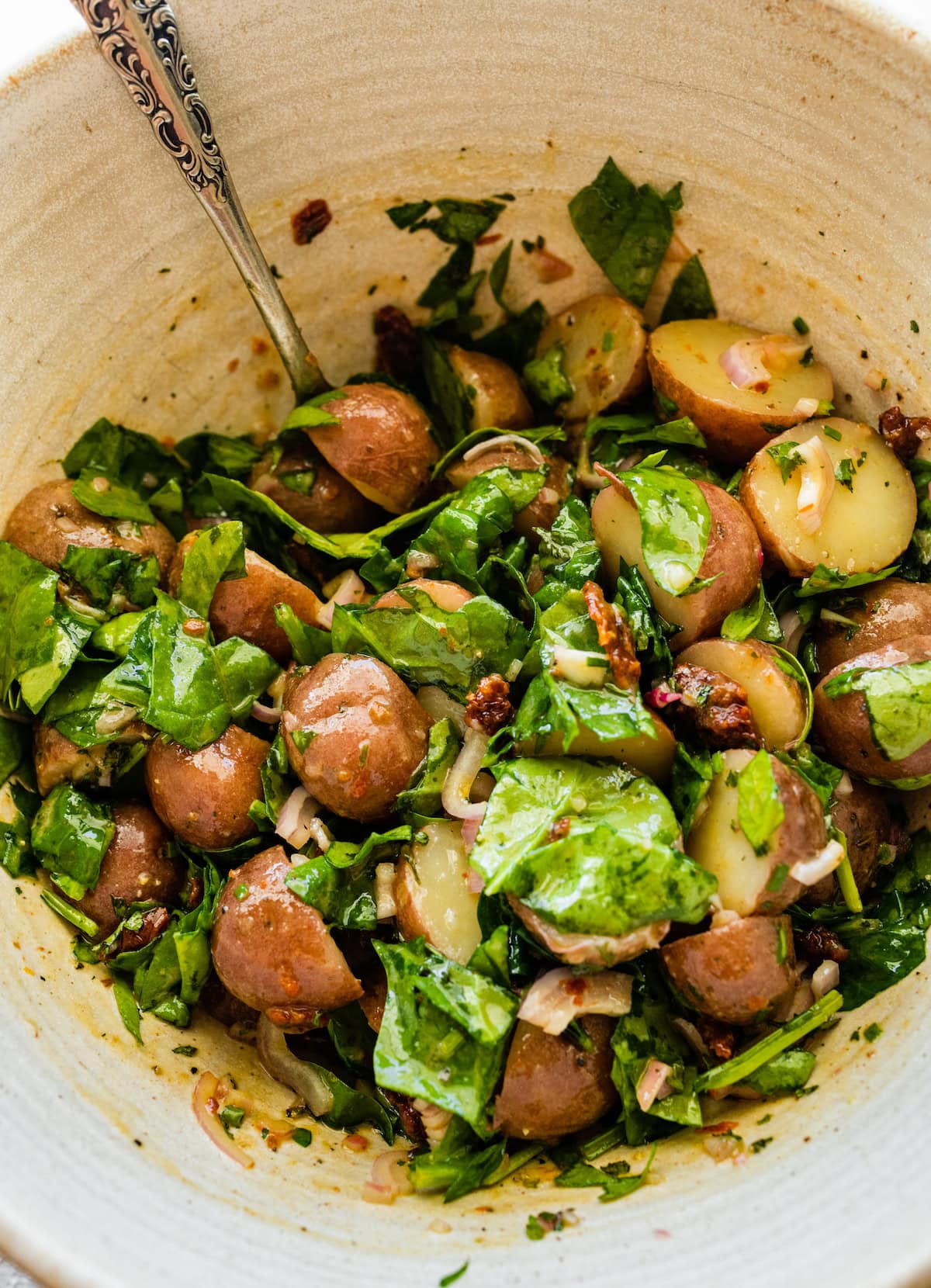 A large bowl with small cooked potatoes, fresh herbs, spinach, and sun dried tomatoes.