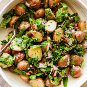 A large bowl with cooked small potatoes, spinach, sun dried tomatoes, fresh herbs, and a metal spoon.