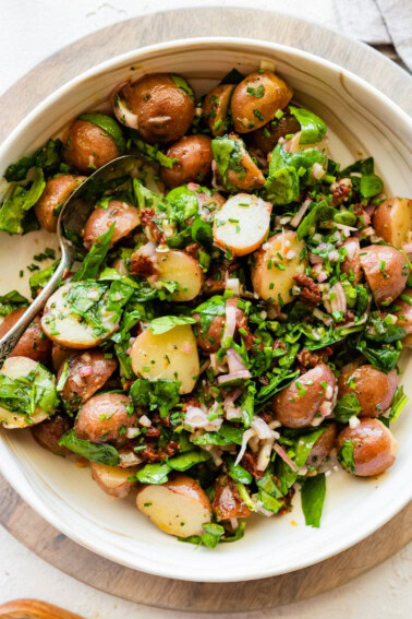 A large bowl with cooked small potatoes, spinach, sun dried tomatoes, fresh herbs, and a metal spoon.