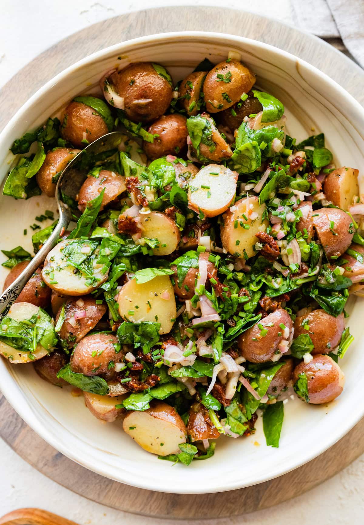 A large bowl with cooked small potatoes, spinach, sun dried tomatoes, fresh herbs, and a metal spoon.
