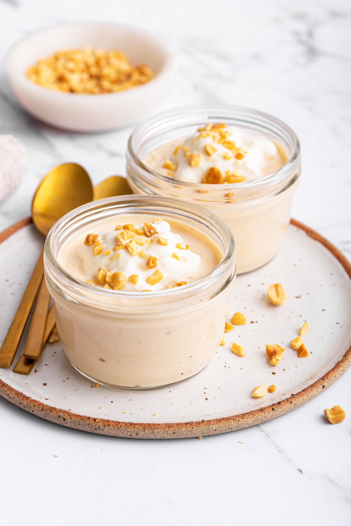 Two glass containers of peanut butter protein pudding on a plate. The pudding is topped with yogurt and crushed peanuts.