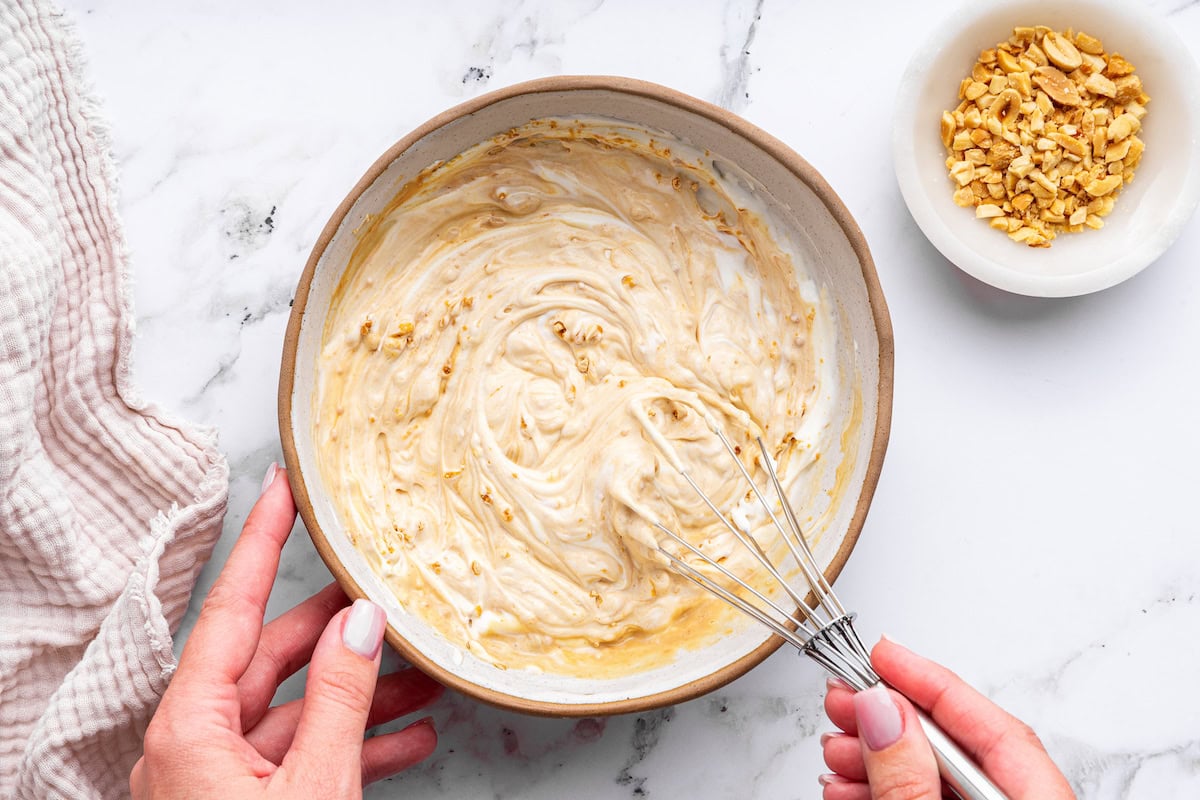 A woman's hand uses a metal whisk to combine ingredients for peanut butter protein pudding in a large mixing bowl.