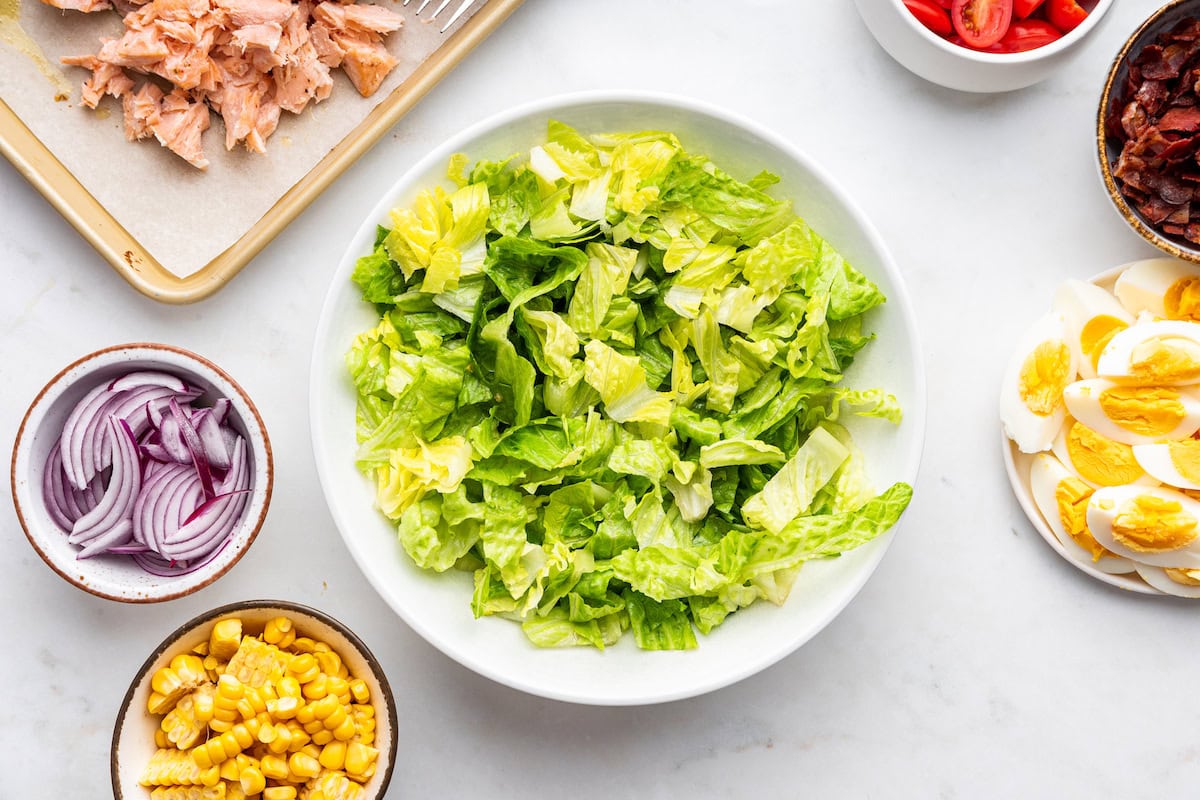 A small white bowl of lettuce with other ingredients near like red onions, corn, and hard boiled eggs.