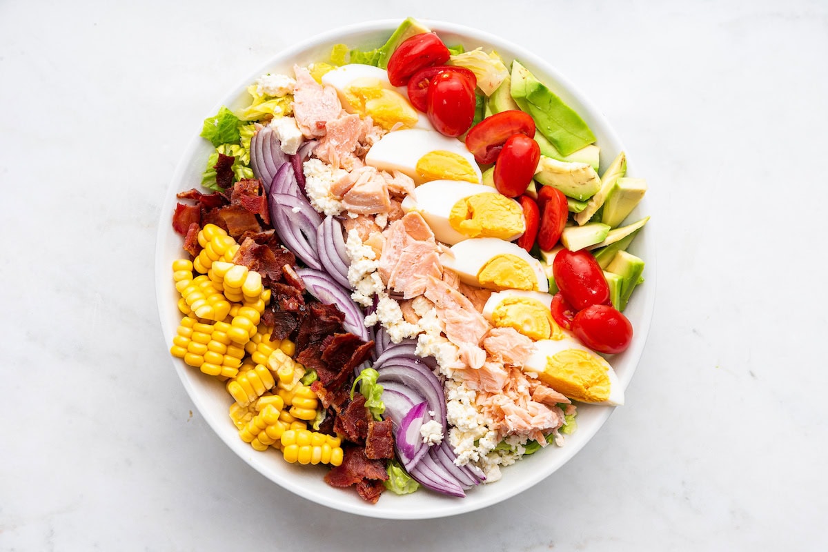 A large bowl of a salmon cobb salad with cooked salmon, feta cheese, red onion, bacon, cherry tomatoes, hard boiled eggs, corn, avocado, and lettuce.