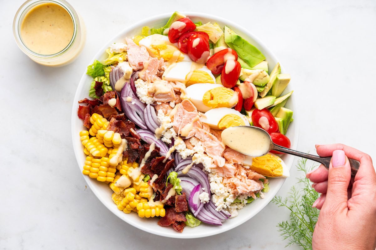 A womans hand uses a spoon to drizzle a honey mustard dressing on a salmon cobb salad.