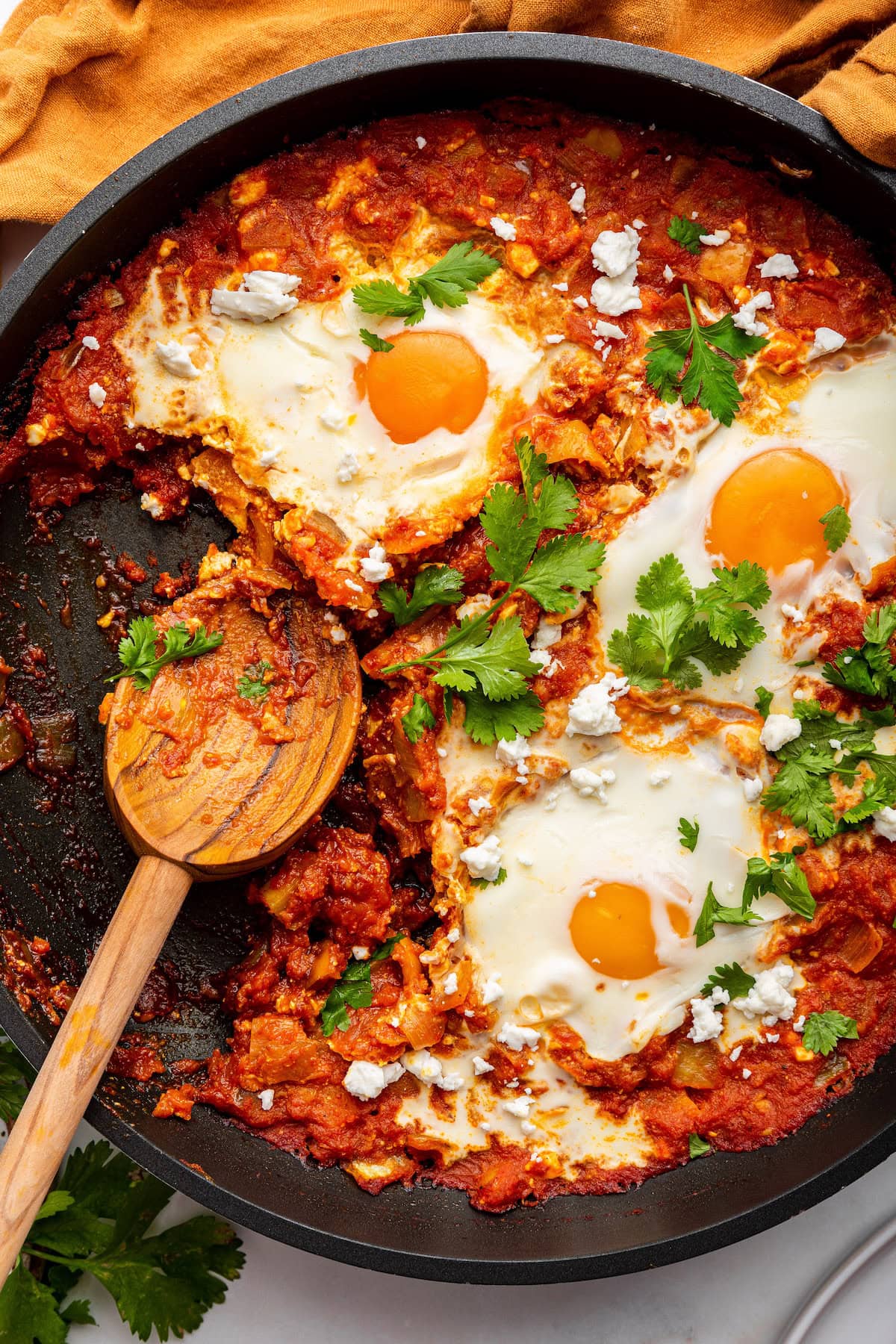 A cast iron skillet with shakshuka. It contains poached eggs in a tomato based sauce and is topped with crumbled feta cheese and fresh herbs.