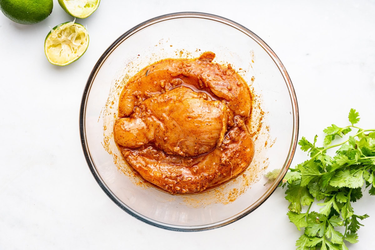 Raw chicken marinating in a large glass bowl of oil, garlic, and spices.