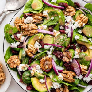 A large serving bowl with fresh spinach, avocado slices, red onion, walnuts, dried cranberries, feta cheese, cucumber, and a homemade dressing.