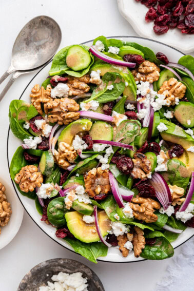 A large serving bowl with fresh spinach, avocado slices, red onion, walnuts, dried cranberries, feta cheese, cucumber, and a homemade dressing.