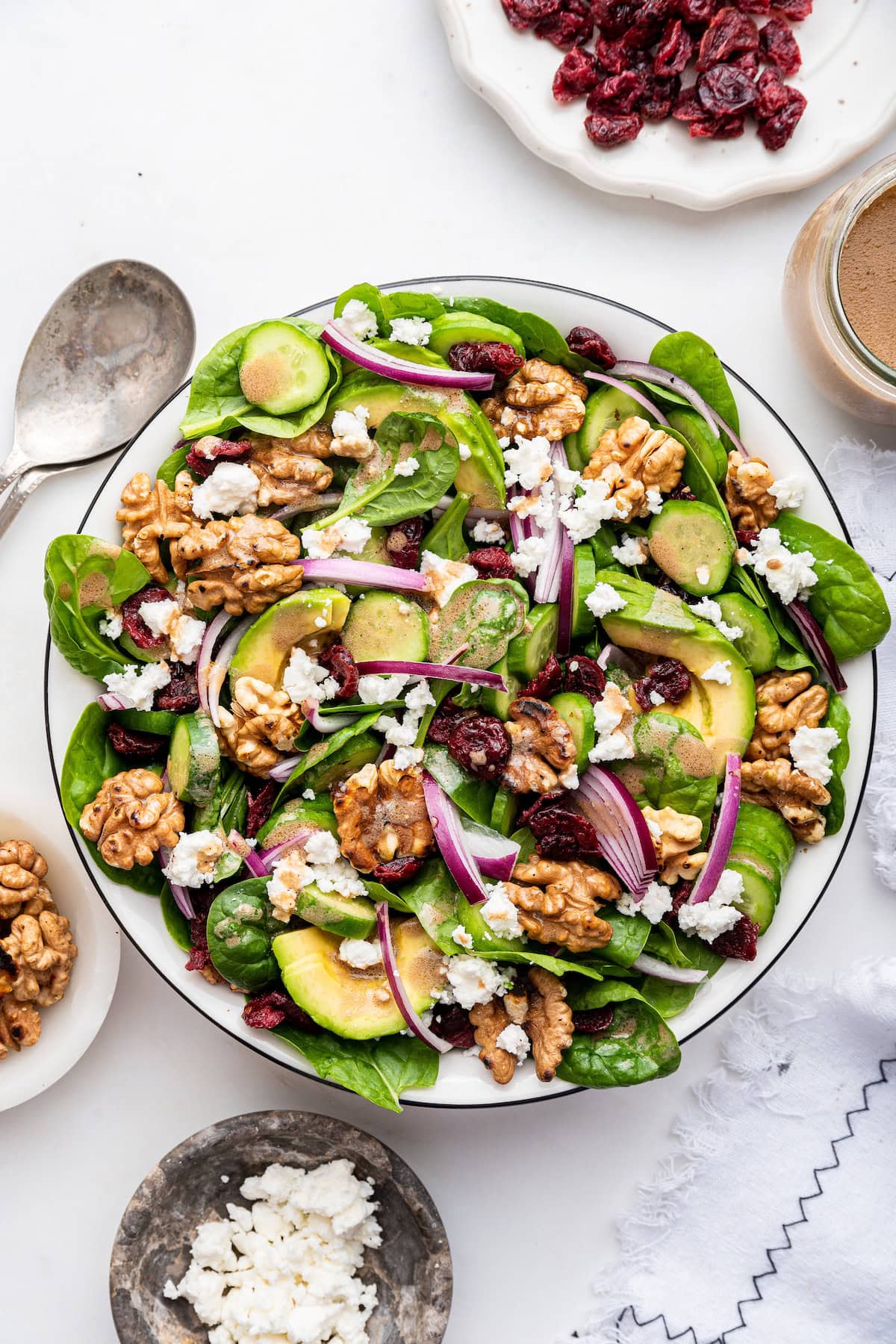A large bowl with fresh spinach, red onion slices, cucumber slices, feta cheese, walnuts, dried cranberries, avocado slices, and a drizzle of dressing.