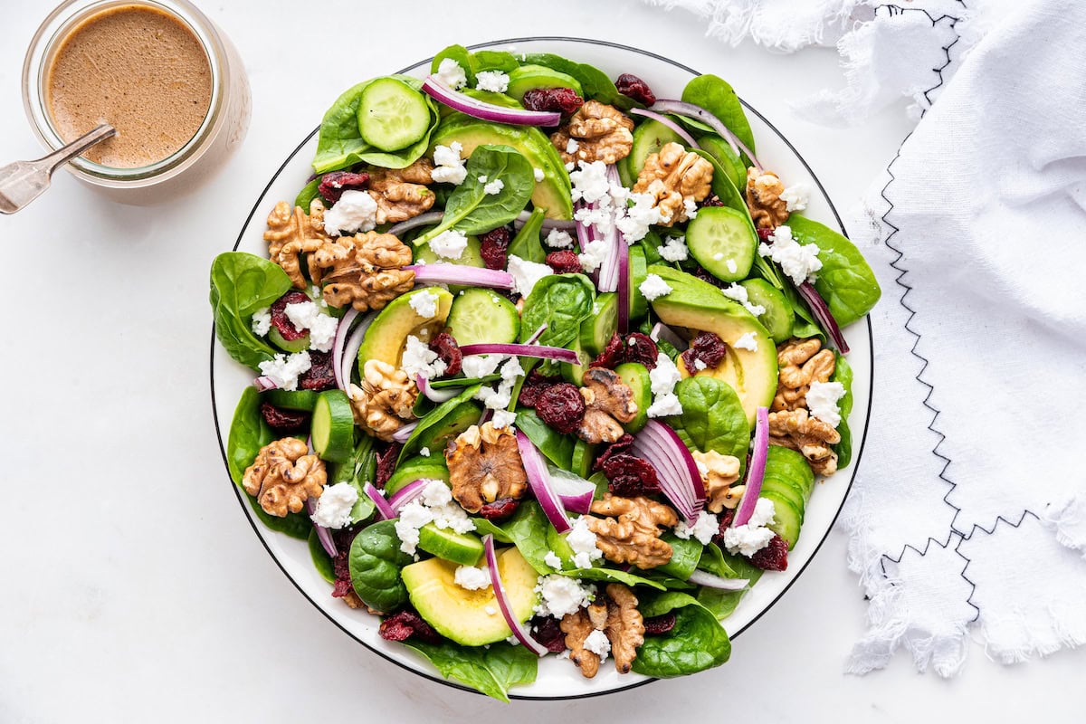 A large serving of fresh spinach, red onion, cucumber slices, red onion, avocado slices, walnuts, dried cranberries, and feta cheese.