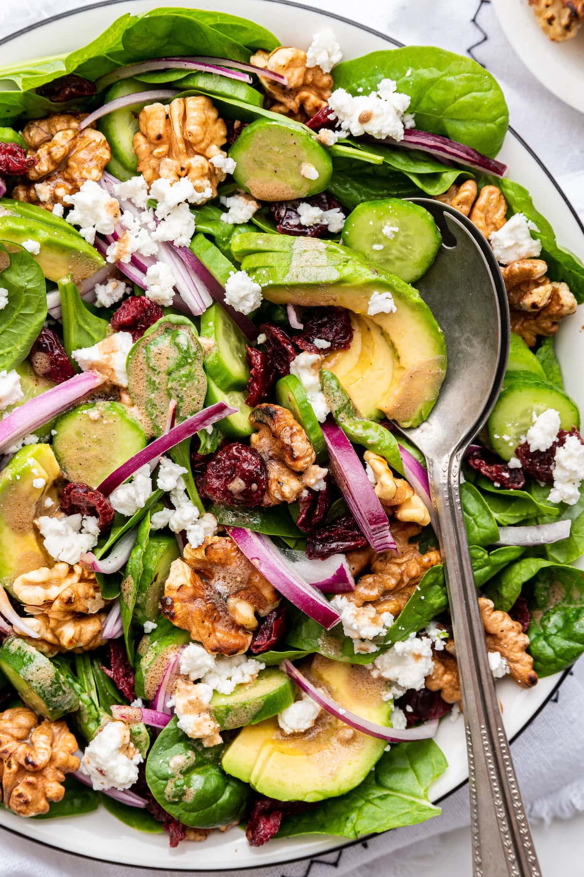 A bowl with a spinach salad with walnuts, dried cranberries, feta cheese, red onion, avocado slices, cucumber slices, and a metal spoon.