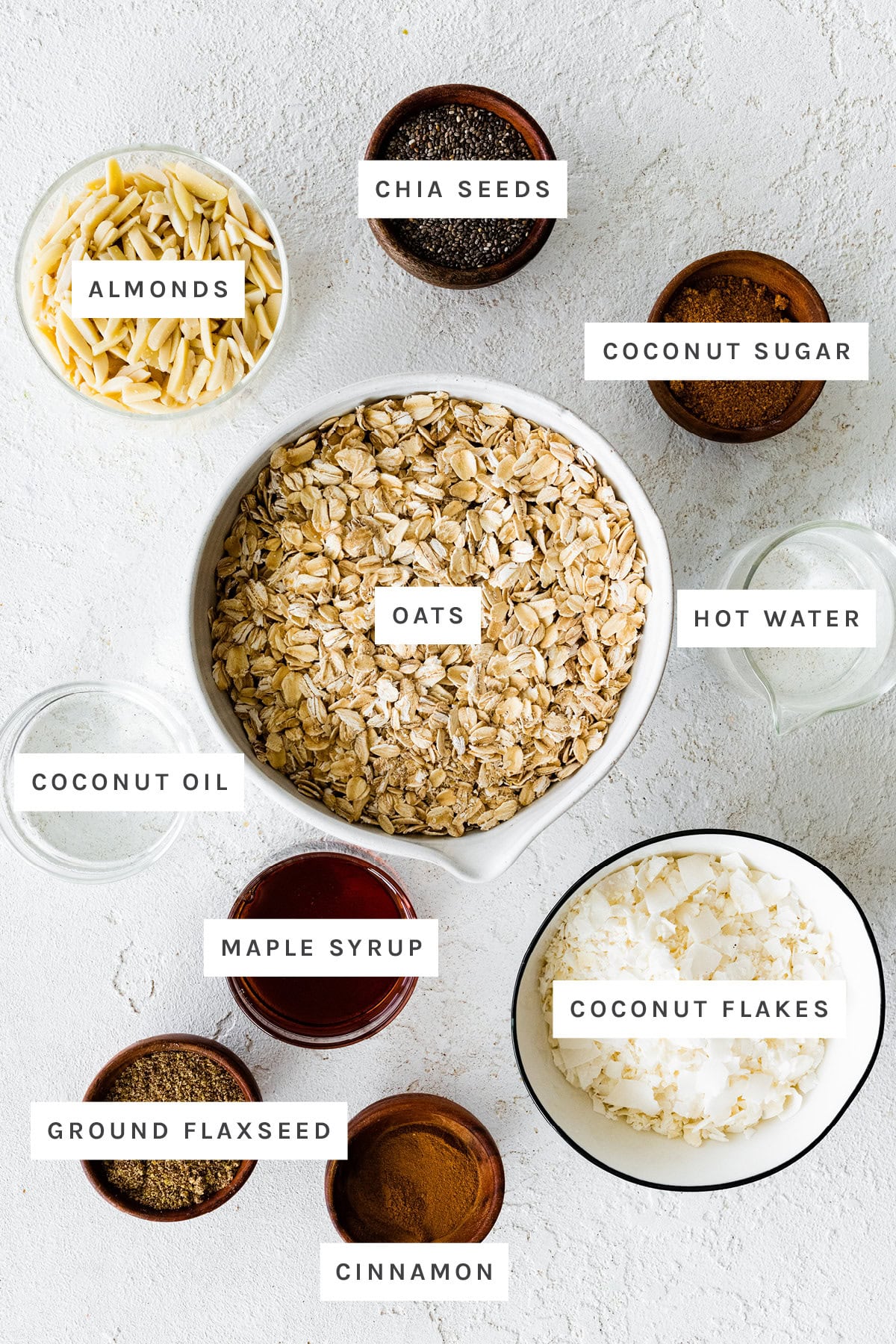 Ingredients measured out to make Coconut Granola: almonds, chia seeds, coconut sugar, oats, hot water, coconut oil, maple syrup, coconut flakes, ground flaxseed and cinnamon.