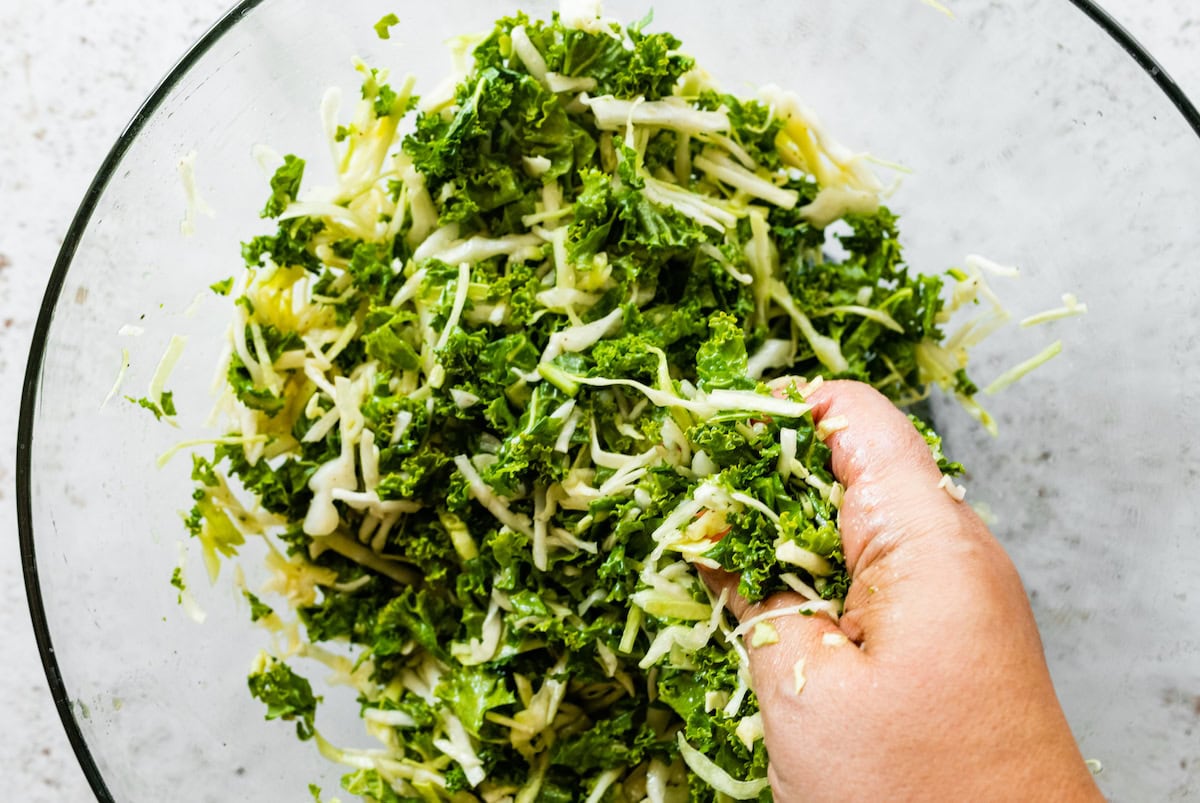 A hand massaging kale and cabbage in a large glass bowl.