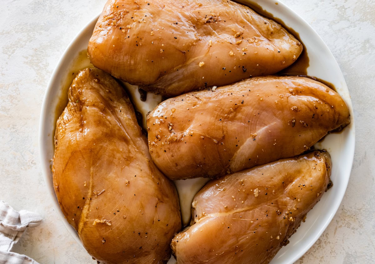 Four raw and marinated chicken breasts on a large plate.