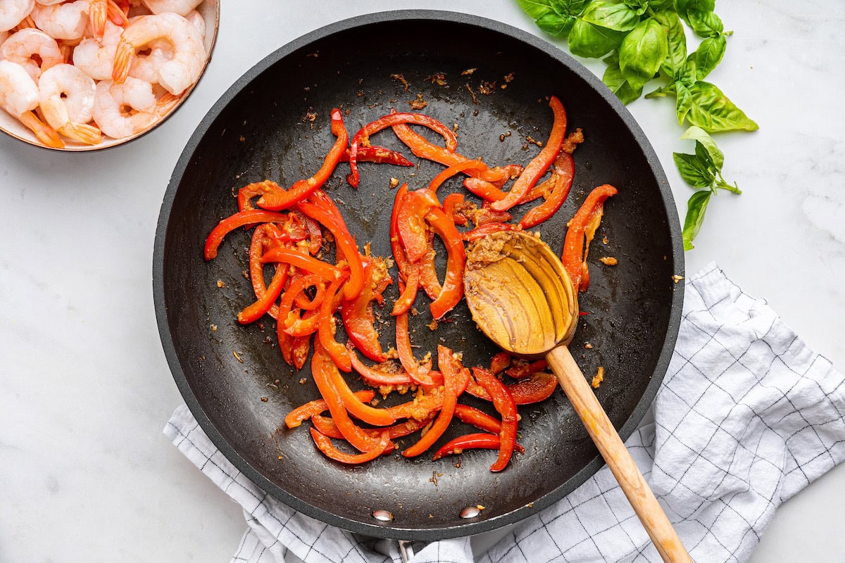 A skillet with red bell pepper and a wooden spoon. There is a bowl of raw shrimp to the side as well as fresh basil.