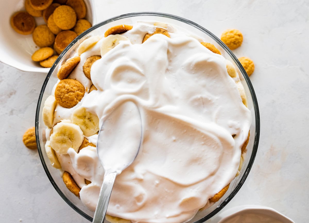 A spoon spreading coco whip on top of layers of fresh banana slices and vanilla wafer cookies.
