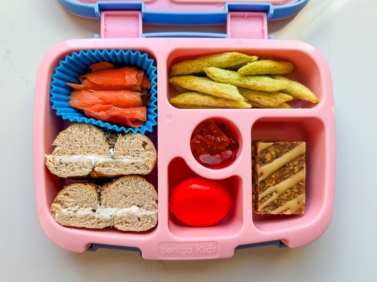 A Bentgo kids lunchbox packed with a mini bagel, smoked salmon, harvest snaps, a babybel cheese, a granola bar and gummies.