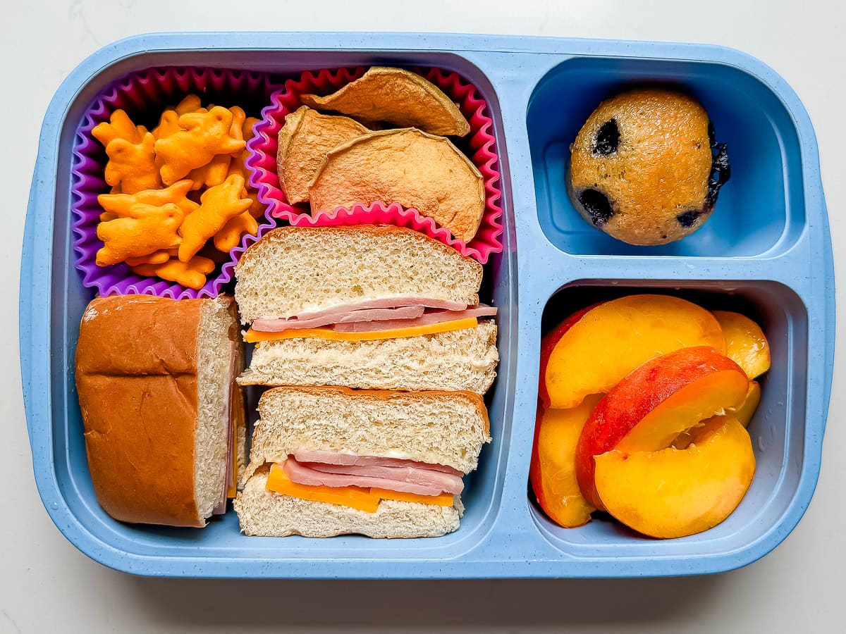 A kids lunch box with ham and cheese sliders, bunny cheese crackers, dried apple chips, a mini blueberry muffin and peaches.