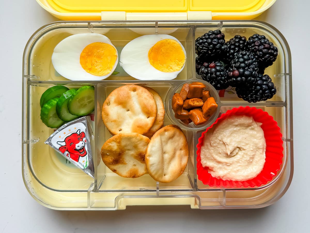 A kids lunch box packed with hummus and pita chips, a laughing cow cheese wedge, cucumbers, a hard boiled egg, blackberries and natural gummies.
