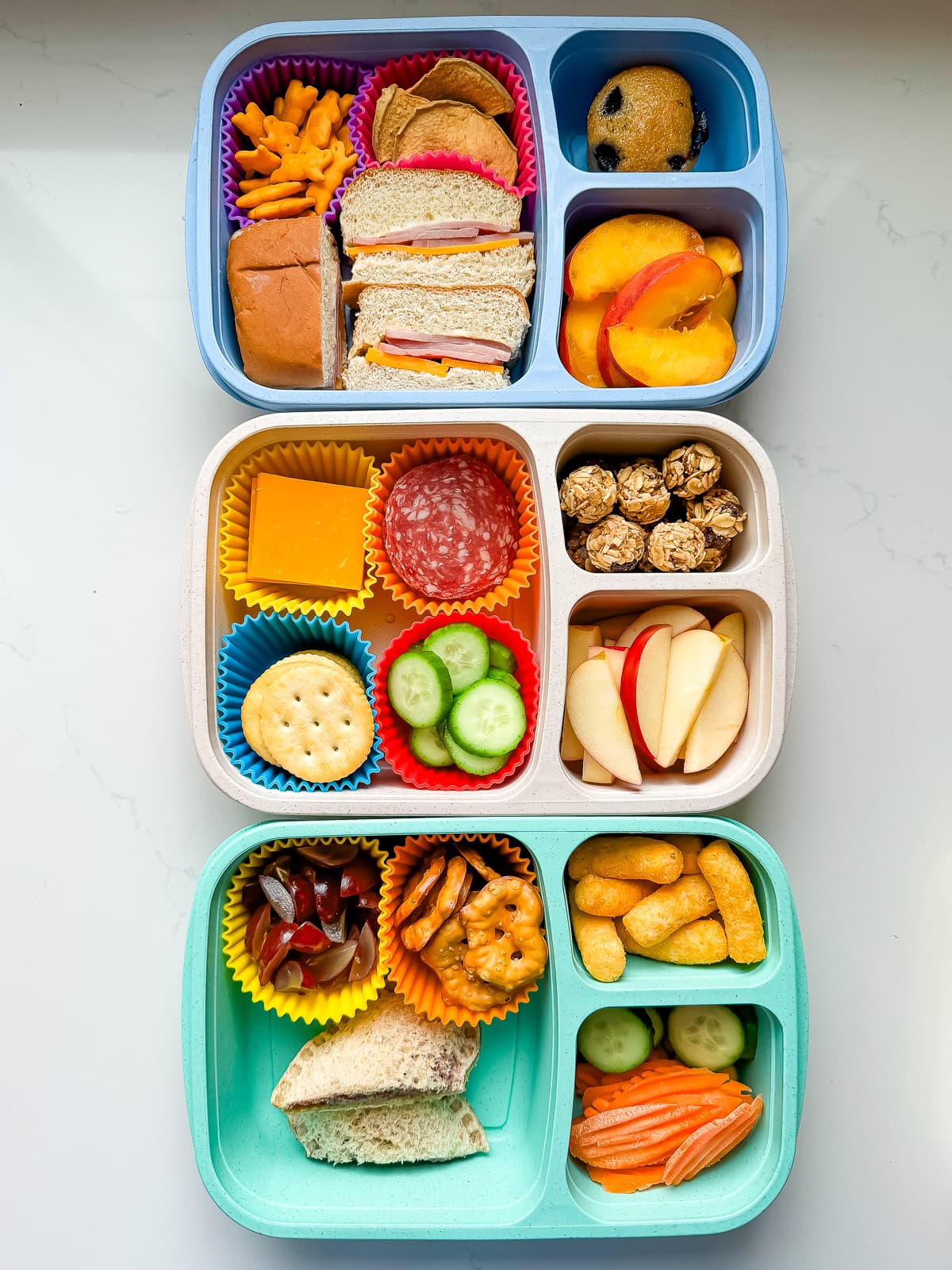 A white countertop with three school lunches packed in colorful lunch containers.