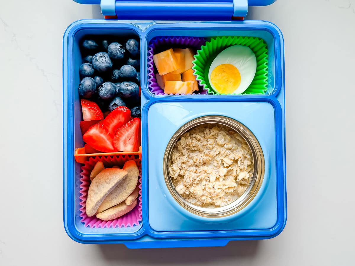 An OmieBox lunch box packed with overnight oats, freeze dried mango, strawberries, blueberries, cheese cubes and a hard boiled egg.