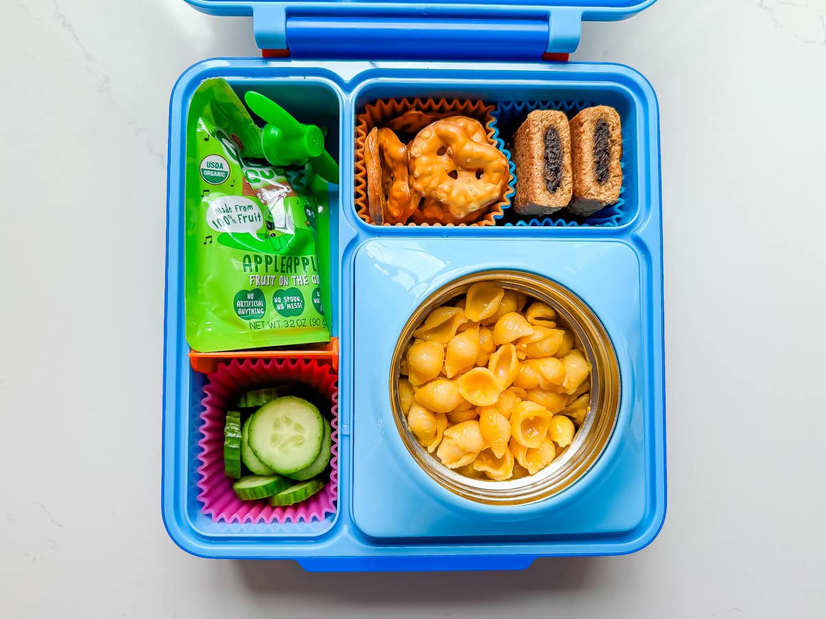 An OmieBox Kid's lunchbox with mac and cheese, cucumbers, an applesauce pouch, pretzel things and a fig bar.