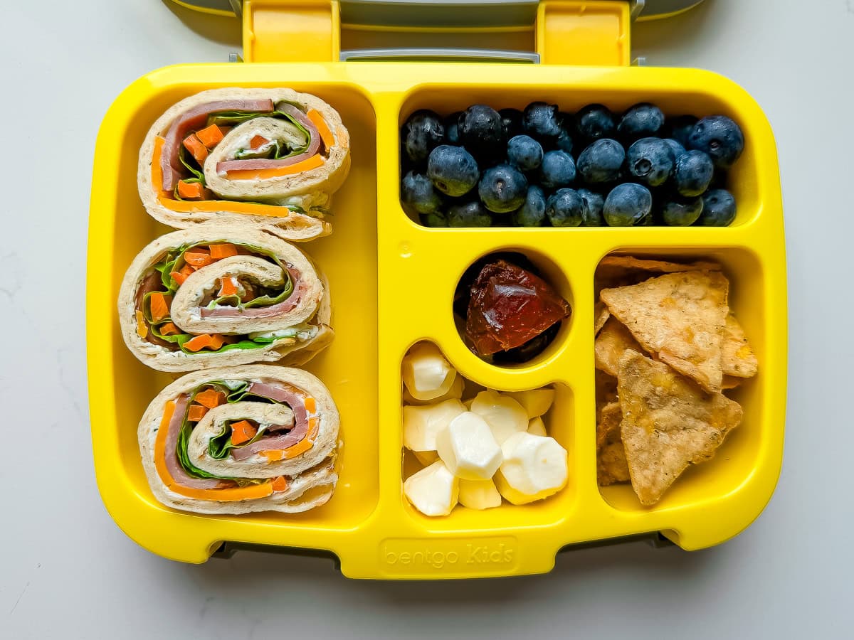 A yellow Bentgo lunchbox packed with pinwheel sandwiches, blueberries, chips, mini mozzarella balls and dates.