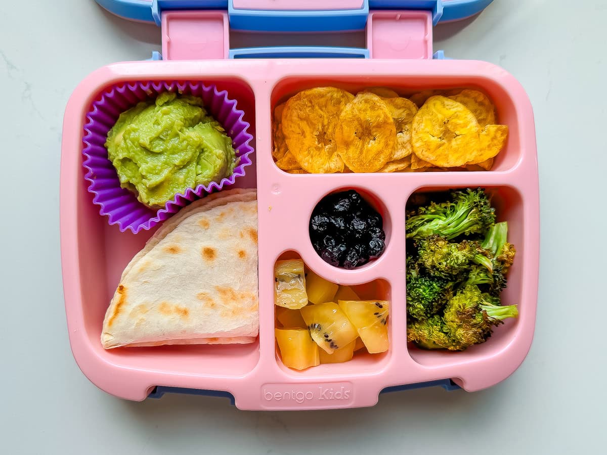 A pink Bentgo lunchbox packed with a quesadilla, guacamole, plantain chips, broccoli, kiwi and dried blueberries.