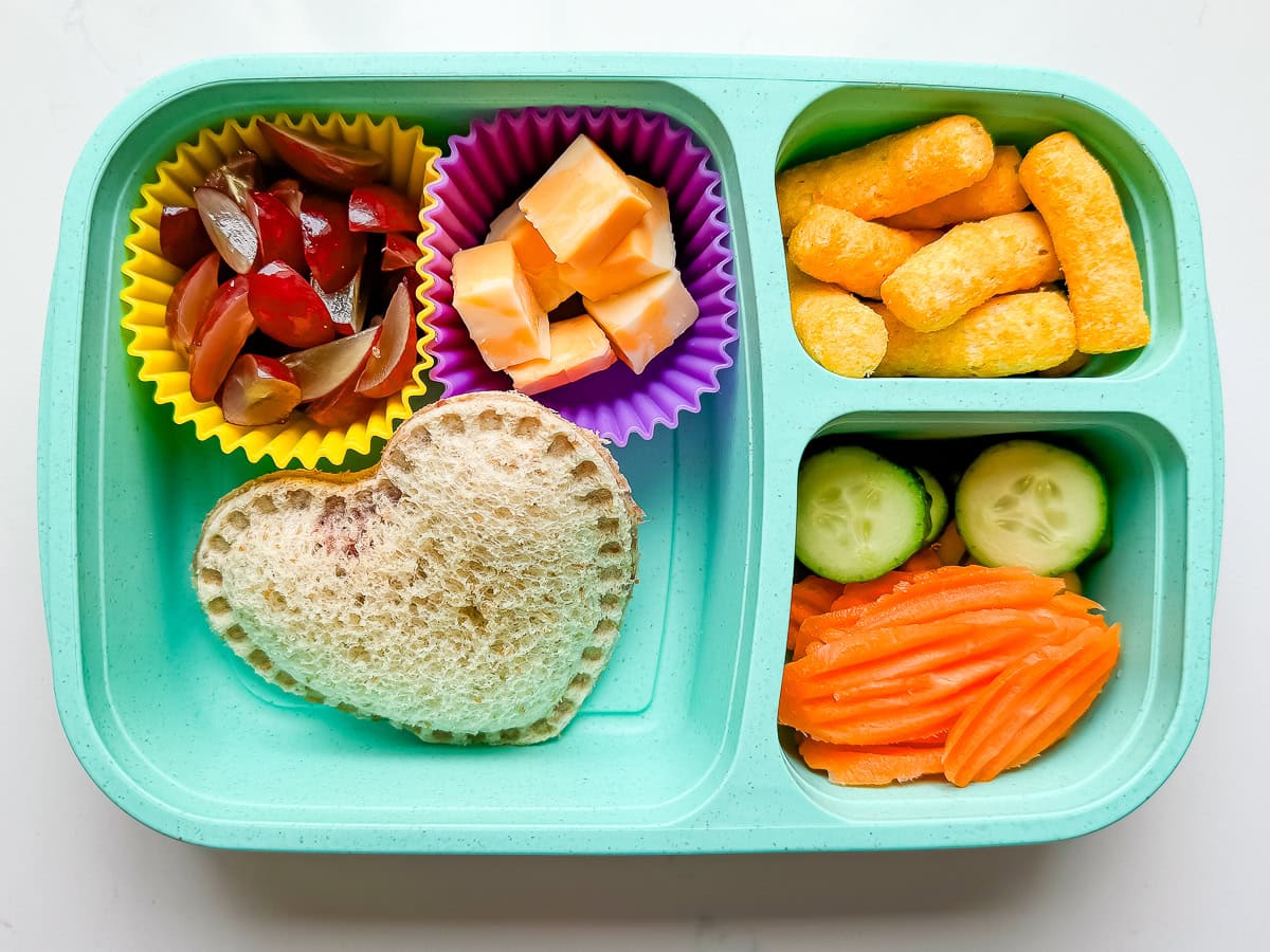 A kids lunch box with a heart shaped DIY uncrustable, grapes, cheese cubes, cheese puffs, carrot chips and cucumbers.