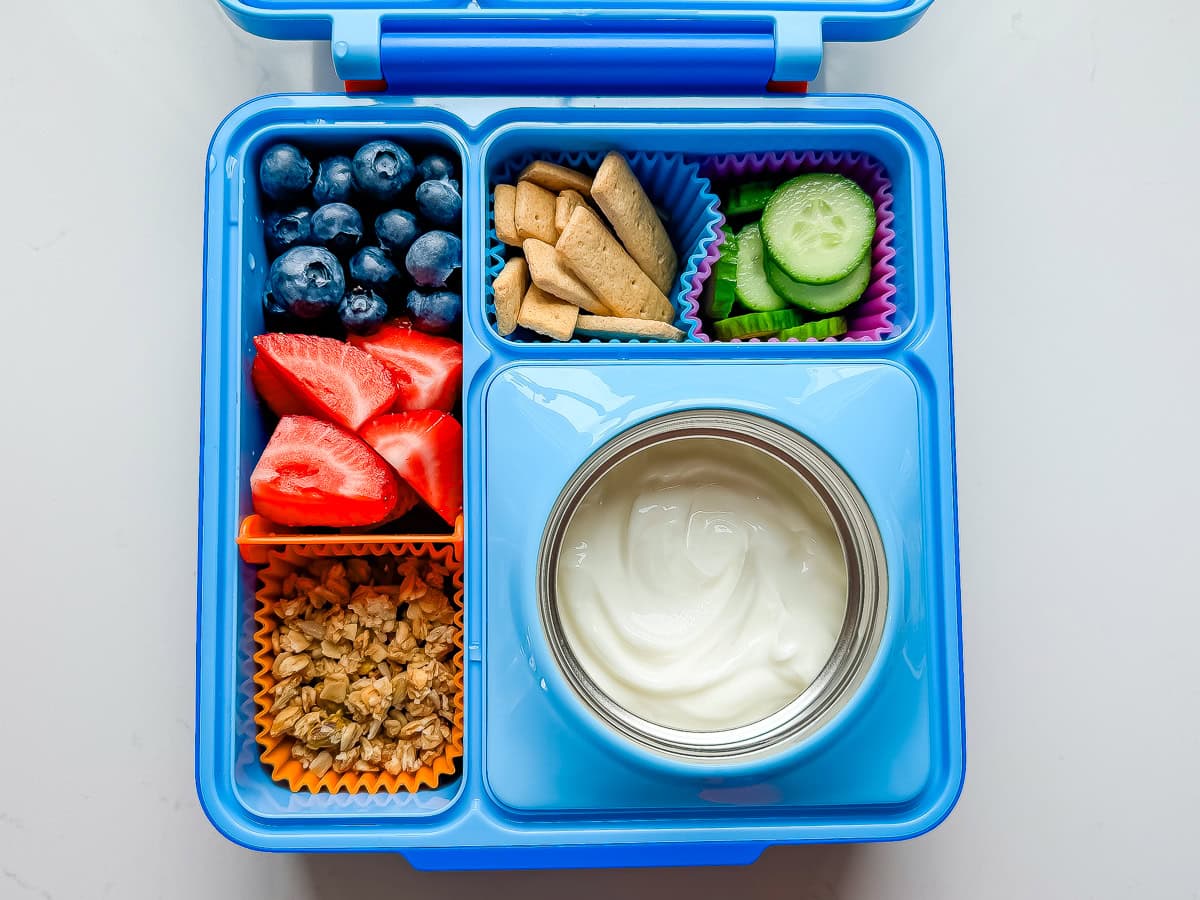 An OmieBox lunchbox packed with Greek yogurt, granola, strawberries, blueberries, graham crackers and cucumbers.