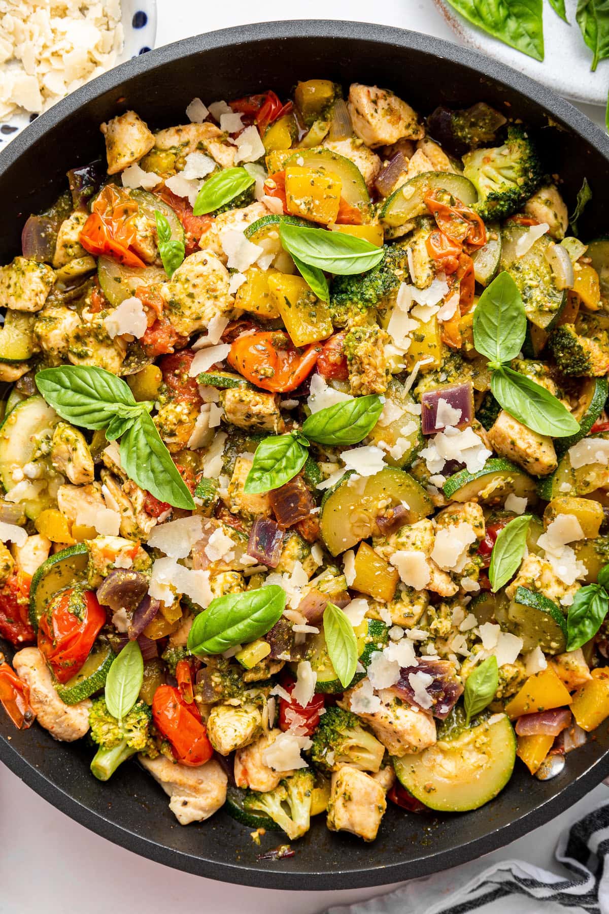 A large skillet with cooked pesto chicken with multiple colorful vegetables and topped with fresh basil leaves.