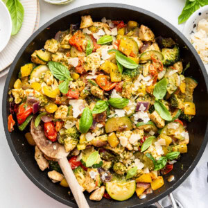 A large skillet with cooked pesto chicken, squash, cauliflower, cherry tomatoes, and fresh basil leaves.