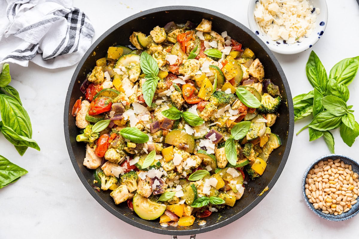 A large skillet with cooked chicken, broccoli, squash, cherry tomatoes, cauliflower, pesto, and fresh basil leaves.