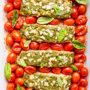 Four salmon filets covered in pesto and surrounded by cherry tomatoes in a baking dish.