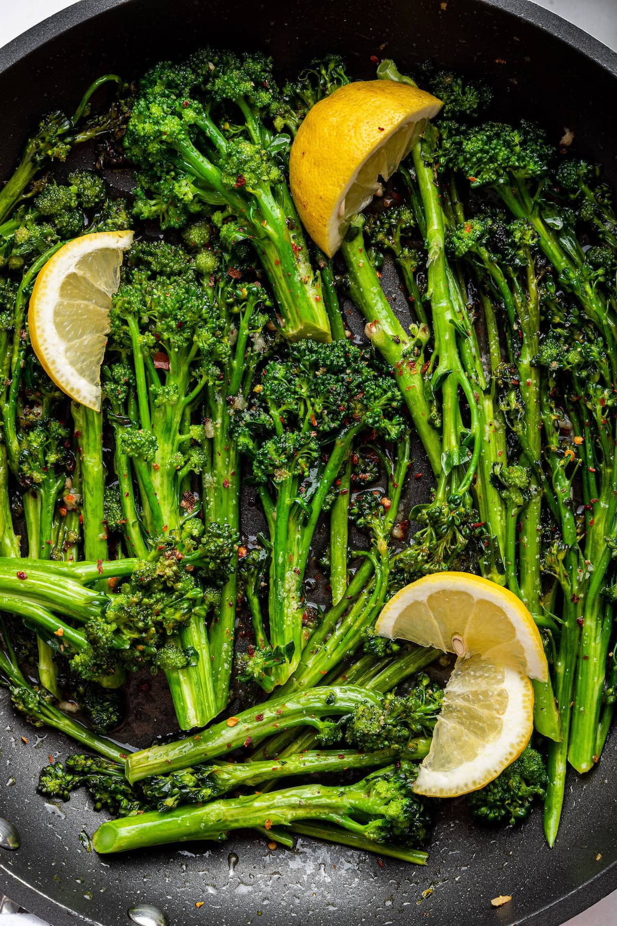 A pan with seasoned and cooked broccolini with fresh lemon wedges.